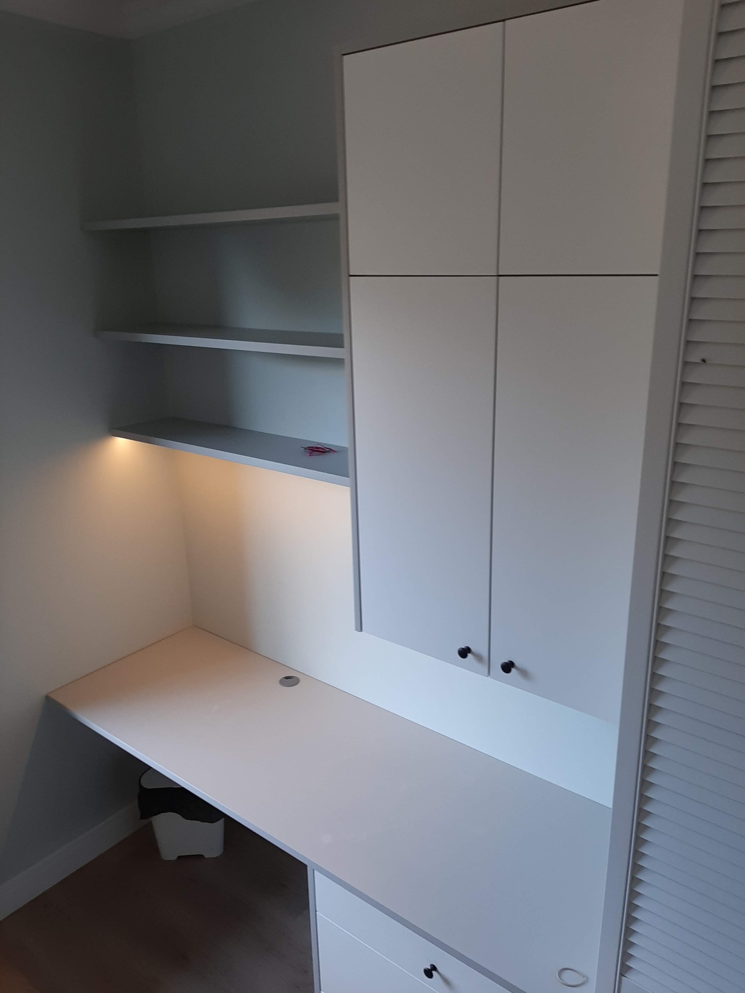 Białe biurko wnękowe z szafkami wiszącymi i oświetleniem LED pod półkami, minimalistyczny design.