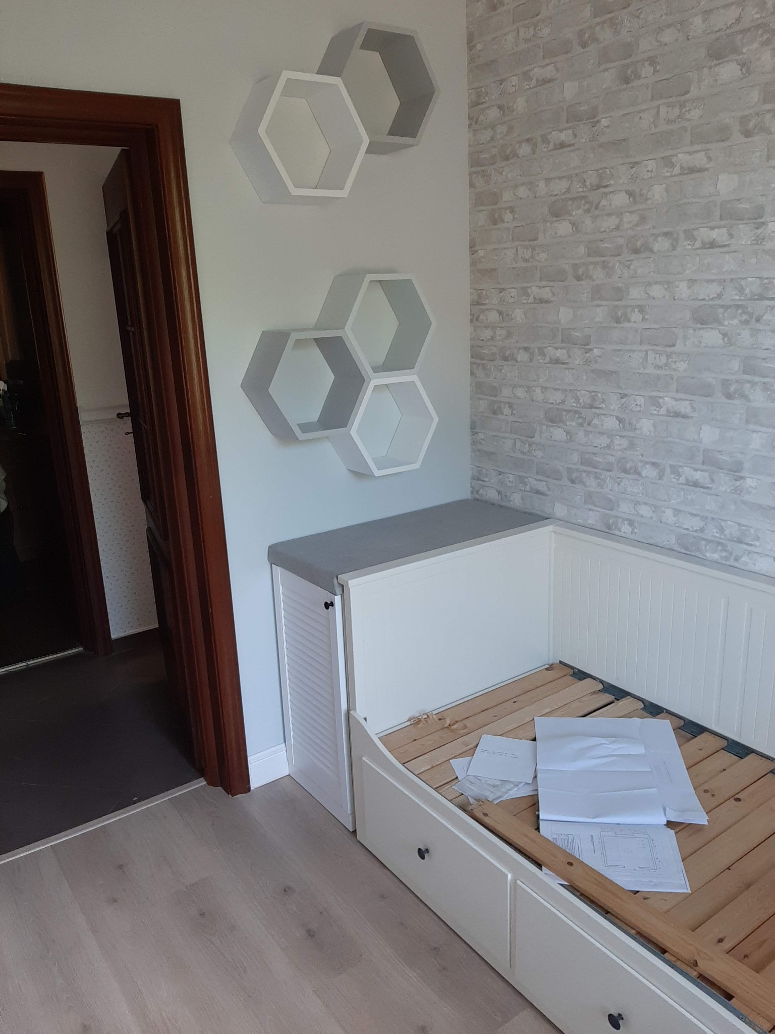 Biała zabudowa meblowa z szufladami i siedziskiem pod ścianą z cegły, uzupełniona o dekoracyjne sześciokątne półki ścienne, widoczne plany na drewnianym stelażu.