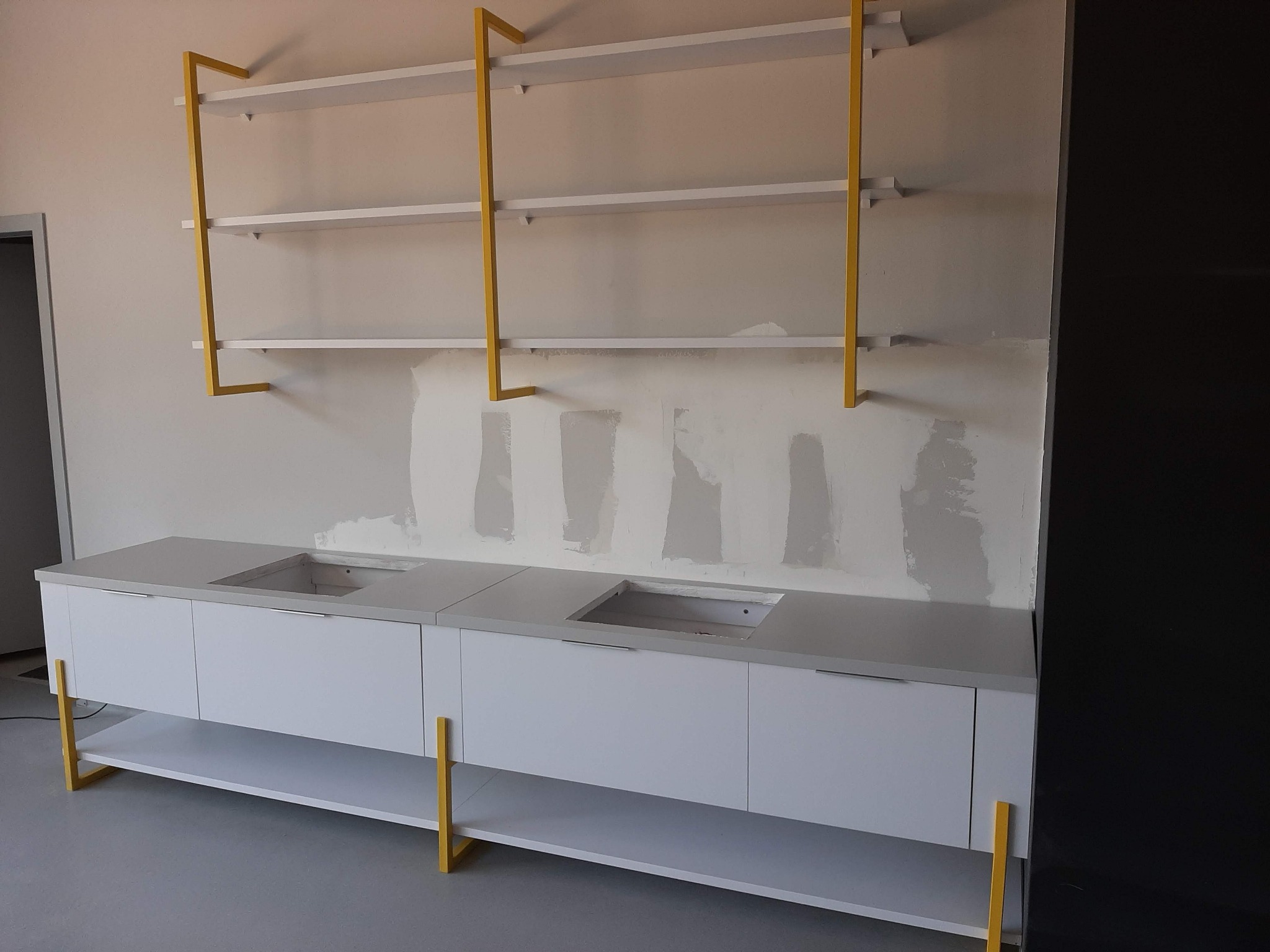 Biała szafka łazienkowa z szarym blatem i żółtymi elementami konstrukcyjnymi, widoczne otwory na umywalki, w tle ściana w trakcie wykończenia i białe półki z żółtymi wspornikami.