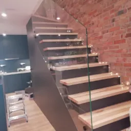 Nowoczesne schody z drewnianymi stopniami, szarą konstrukcją i szklaną balustradą, oświetlone delikatnymi lampkami, na tle ceglanej ściany i fragmentu kuchni z metalową drabinką.