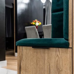 ARTezze - Fragment zabudowy: drewnopodobna szafka z siedziskiem i tapicerowanym panelem ściennym w kolorze butelkowej zieleni, w tle jadalnia z kwiatami.