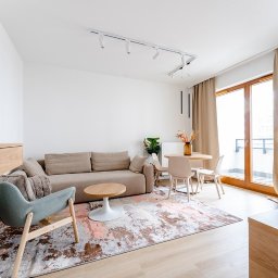 ARTezze - Jasne wnętrze salonu z beżową sofą, okrągłym stolikiem kawowym na białej nodze, turkusowym fotelem na drewnianych nogach, okrągłym stołem jadalnym z czterema krzesłami i wzorzystym dywanem.