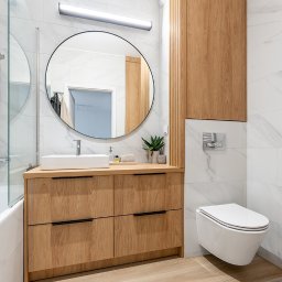 ARTezze - Jasna łazienka z drewnianą szafką pod umywalką z czterema szufladami i okrągłym lustrem, biała toaleta podwieszana, marmurowe płytki na ścianach.