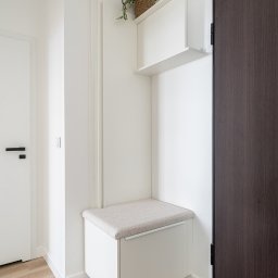ARTezze - Biała zabudowa w przedpokoju z siedziskiem i szafką wiszącą, uzupełniona rośliną w plecionym koszyku, ciemne drzwi po prawej stronie.