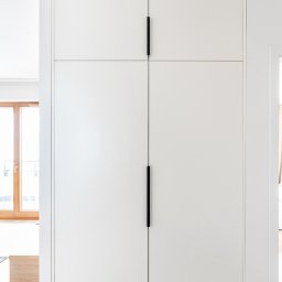 ARTezze - Biała, minimalistyczna szafa wnękowa z czterema drzwiami i długimi, czarnymi uchwytami, widoczny fragment logo firmy w dolnej części.