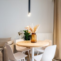 ARTezze - Jasne wnętrze jadalni z okrągłym drewnianym stołem na białej nodze, otoczonym czterema białymi krzesłami. Na stole brązowy wazon z suszonymi kwiatami. W tle sofa, lampa i roślina doniczkowa.