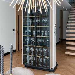 ARTezze - Nowoczesna przeszklona szafa ekspozycyjna z czarnymi ramami, wypełniona różnymi rodzajami kieliszków do wina, podświetlona od wewnątrz, widoczna w eleganckim wnętrzu z parkietem w jodełkę...