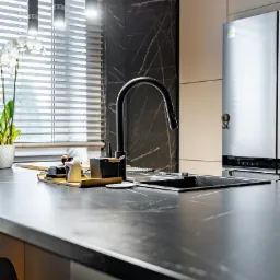 Czarny, marmurkowy blat kuchenny z minimalistycznym zlewem i czarną baterią, oświetlony wiszącymi lampami cylindrycznymi, z widoczną orchideą w tle i fragmentem krzesła barowego.