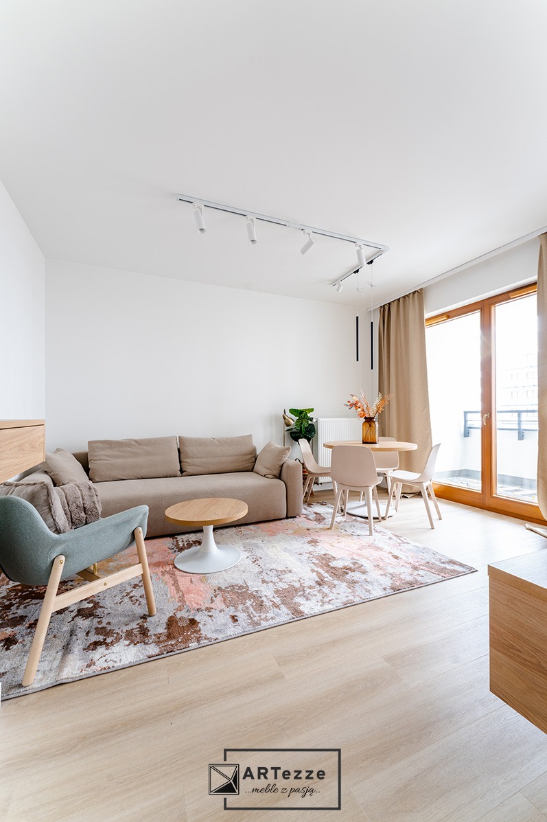 Jasne wnętrze salonu z beżową sofą, okrągłym stolikiem kawowym na białej nodze, turkusowym fotelem na drewnianych nogach, okrągłym stołem jadalnym z czterema krzesłami i wzorzystym dywanem.