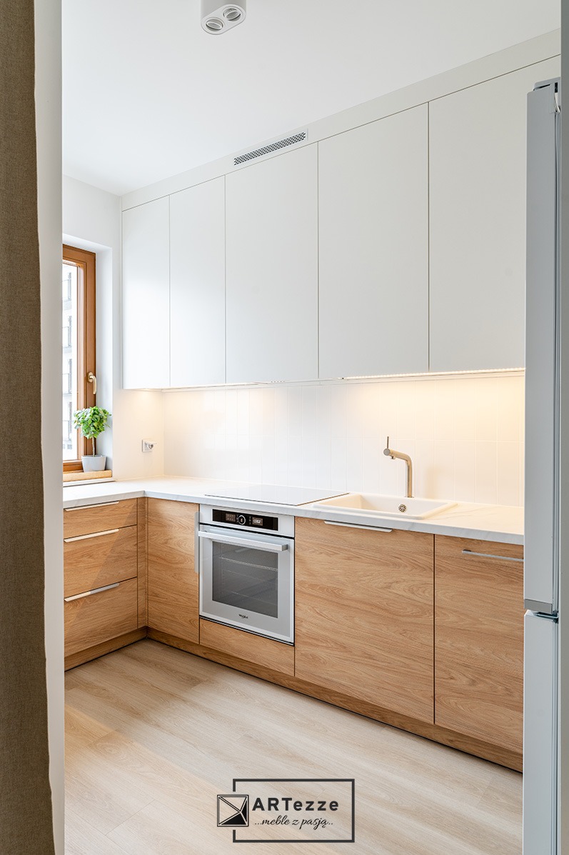 Nowoczesna, jasna kuchnia: dolne szafki z forniru dębowego, białe górne szafki, biały blat, okno z rośliną, oświetlenie LED podszafkowe, zlew z baterią, piekarnik i płyta indukcyjna.