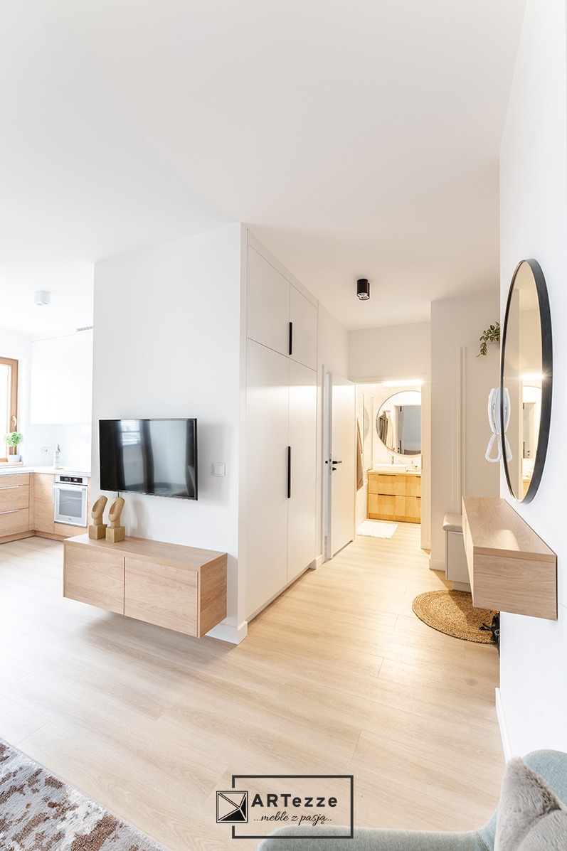 Nowoczesne wnętrze: biała szafa w zabudowie z długimi, czarnymi uchwytami, drewniana szafka RTV pod telewizorem oraz widoczny fragment kuchni z jasnymi, drewnianymi frontami i łazienki z lustrem...