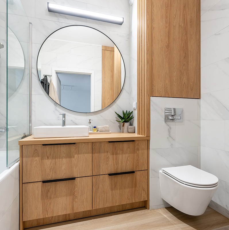 Jasna łazienka z drewnianą szafką pod umywalką z czterema szufladami i okrągłym lustrem, biała toaleta podwieszana, marmurowe płytki na ścianach.