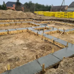 Wylane fundamenty pod budynek mieszkalny, szalunki drewniane wypełnione świeżym betonem, w tle palety z bloczkami Ytong i budynki mieszkalne.