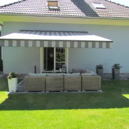 Zadaszony taras przy domu z meblami ogrodowymi z technorattanu, dekoracyjnymi donicami i zadbanym trawnikiem w słoneczny dzień.