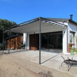 Nowoczesna pergola metalowa z rozsuwanym, jasnym zadaszeniem, przylegająca do białego domu z dużymi przesuwnymi oknami, na betonowym tarasie z okrągłym stolikiem i krzesłami.