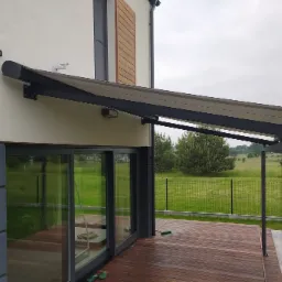 Nowoczesny taras z drewnianą podłogą i zadaszeniem w kolorze antracytowym, przylegający do domu z dużymi przesuwnymi oknami, widok na zielony ogród i metalowe ogrodzenie.