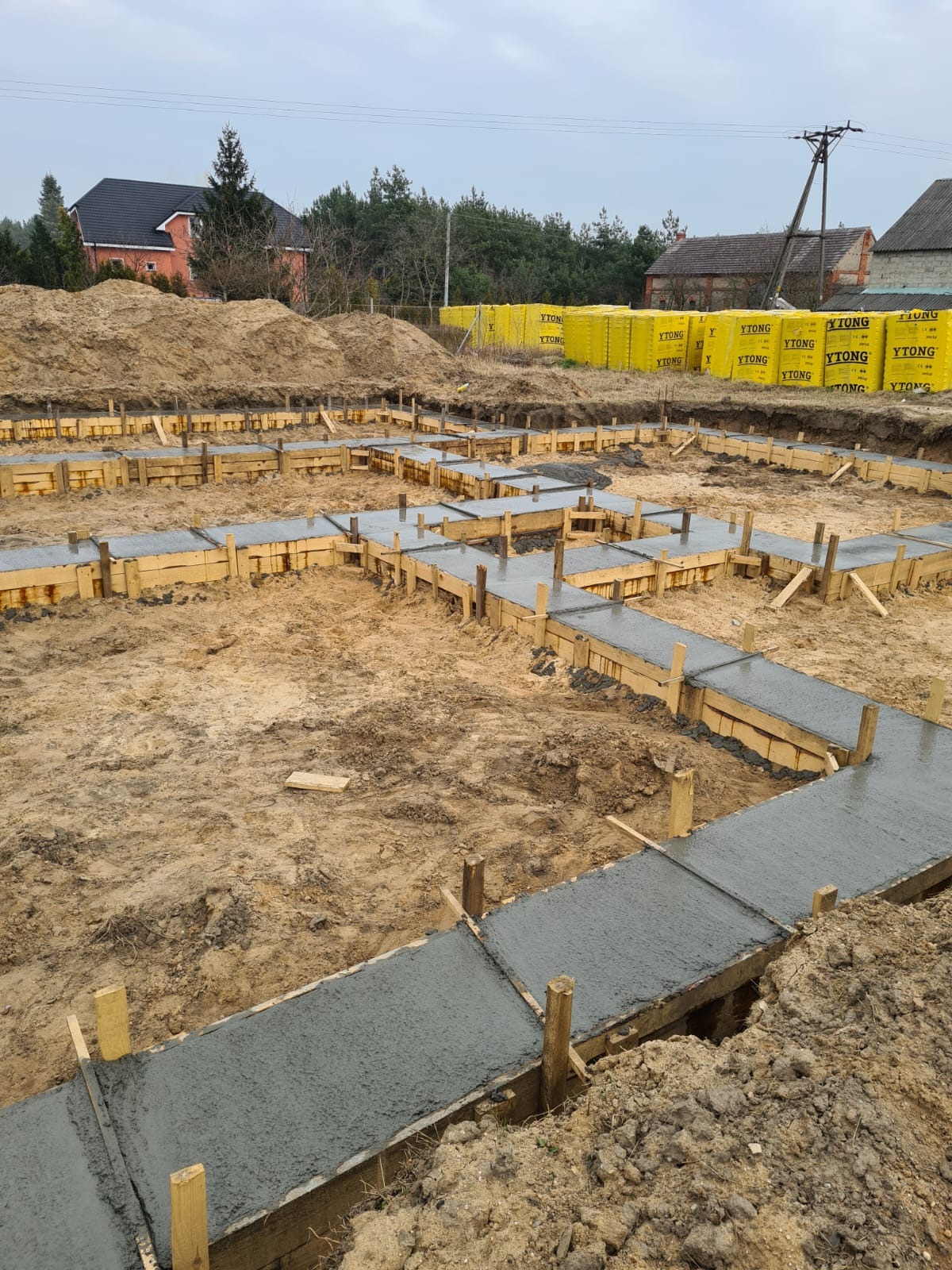 Wylane fundamenty pod budynek mieszkalny, szalunki drewniane wypełnione świeżym betonem, w tle palety z bloczkami Ytong i budynki mieszkalne.