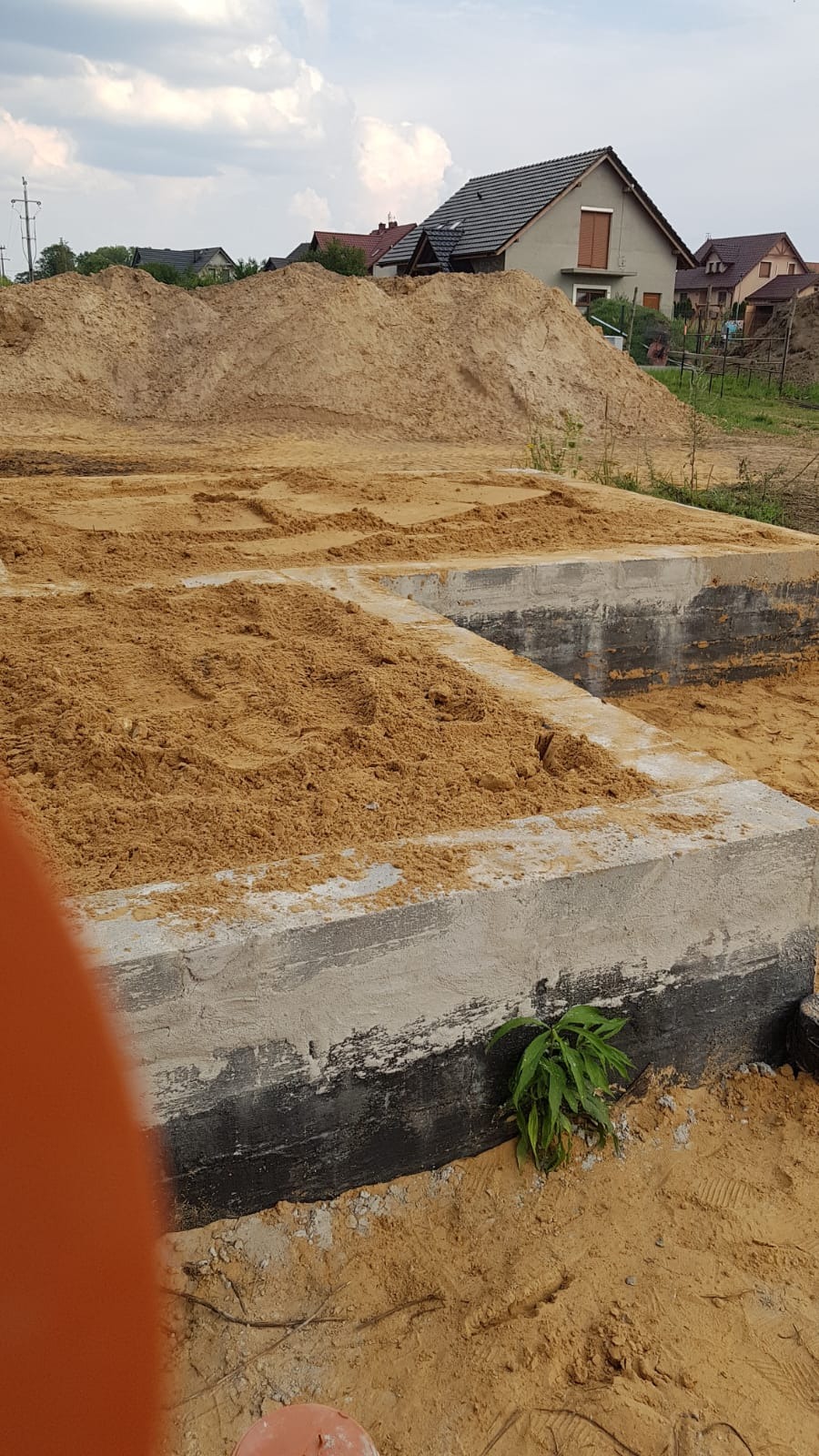 Wylane betonowe fundamenty pod taras lub altanę zasypane piaskiem, widoczne zabezpieczenie przeciwwilgociowe, w tle hałda piasku i domy jednorodzinne.