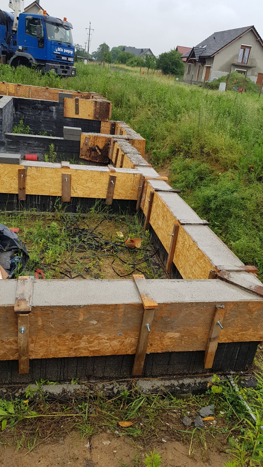Wylany fundament betonowy z deskowaniem na działce z wysoką trawą, w tle dom jednorodzinny i betoniarka.