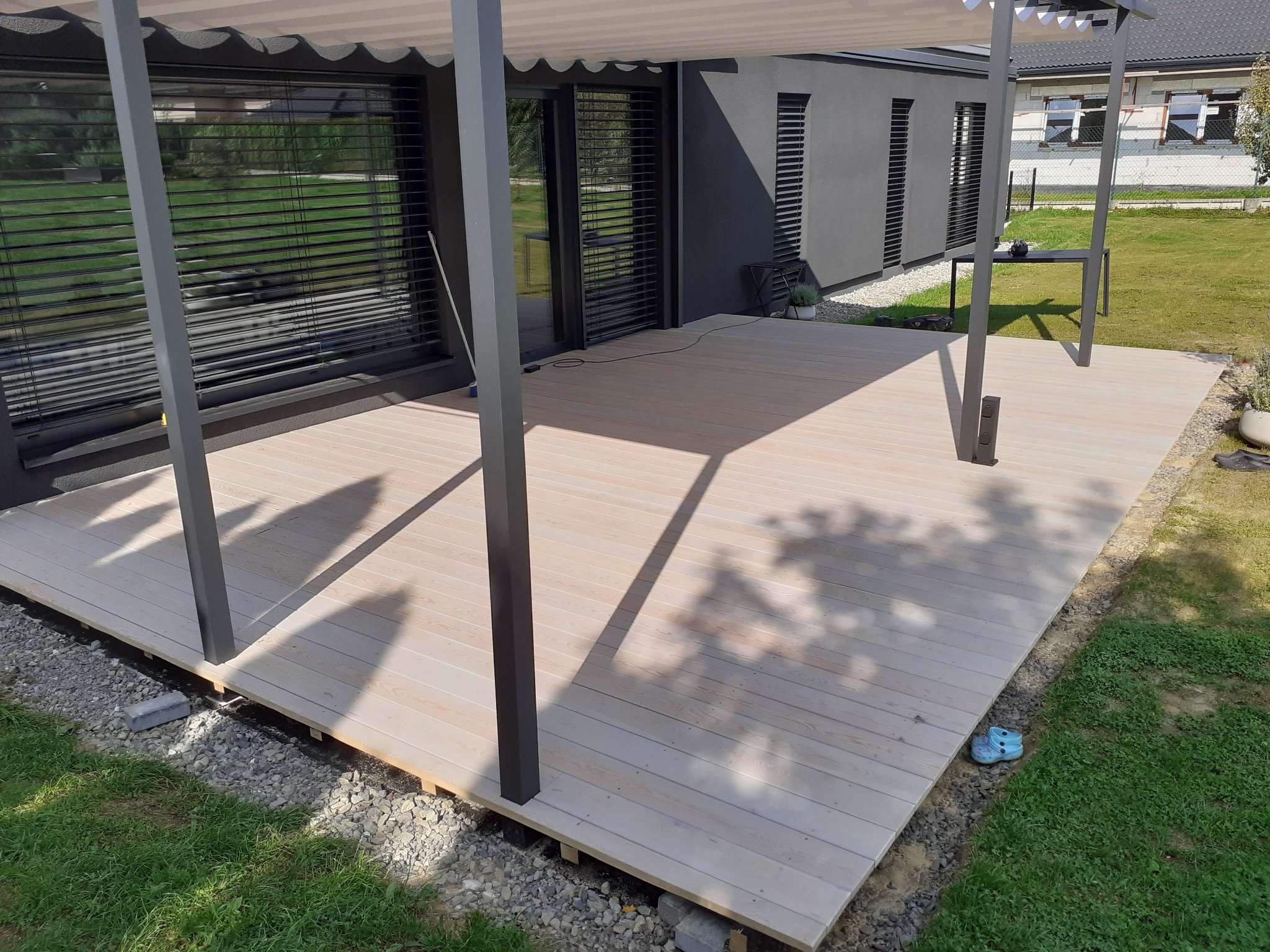 Jasne deski tarasowe pod szarą, nowoczesną pergolą z zadaszeniem, widok na dom z żaluzjami zewnętrznymi, w tle trawnik i budynek w trakcie budowy.