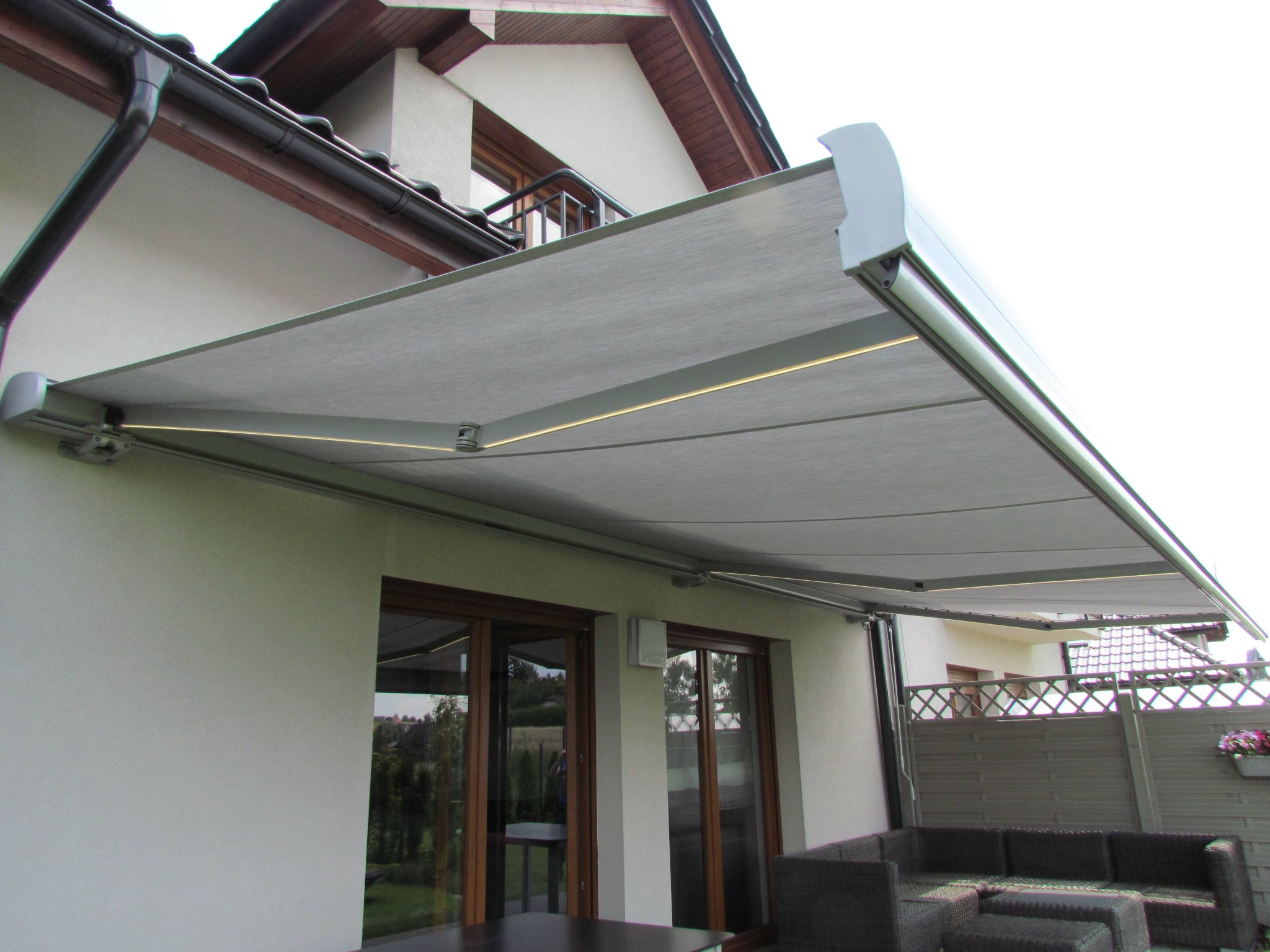 Zewnętrzna, rozkładana markiza z szarej tkaniny, zamontowana nad tarasem przylegającym do domu z brązowymi oknami. Widoczne meble ogrodowe z rattanu.