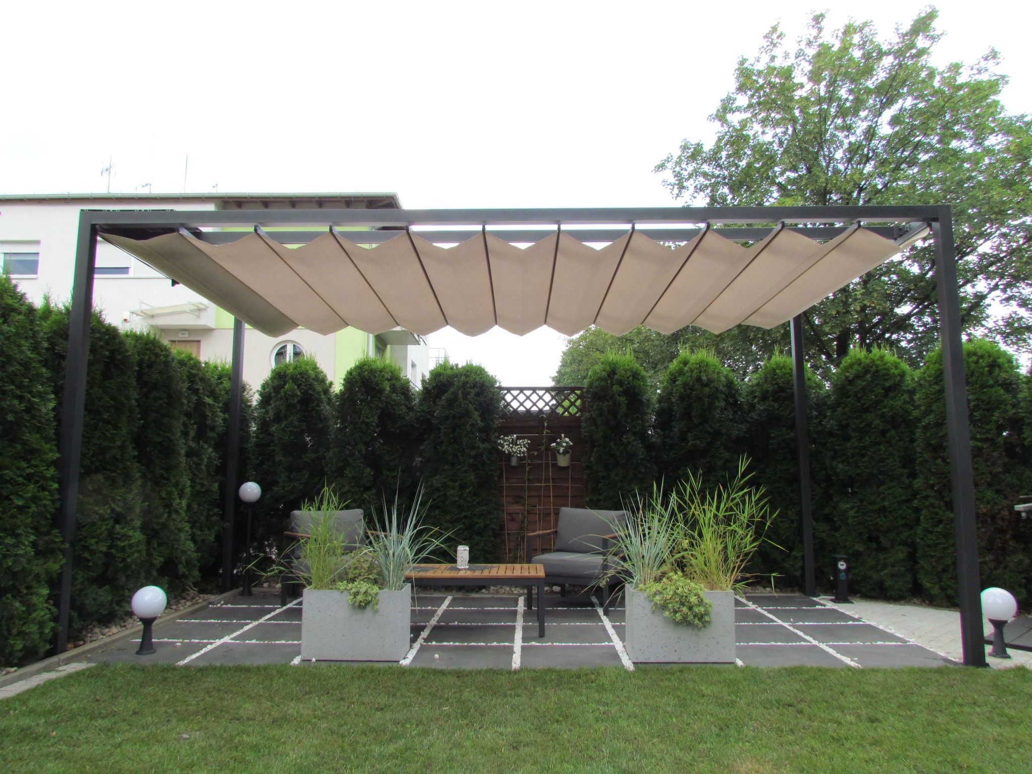 Nowoczesna metalowa konstrukcja z rozsuwanym beżowym zadaszeniem nad strefą relaksu z dwoma fotelami, stolikiem i betonowymi donicami z zielenią, otoczona tujami i oświetlona kulistymi lampami.