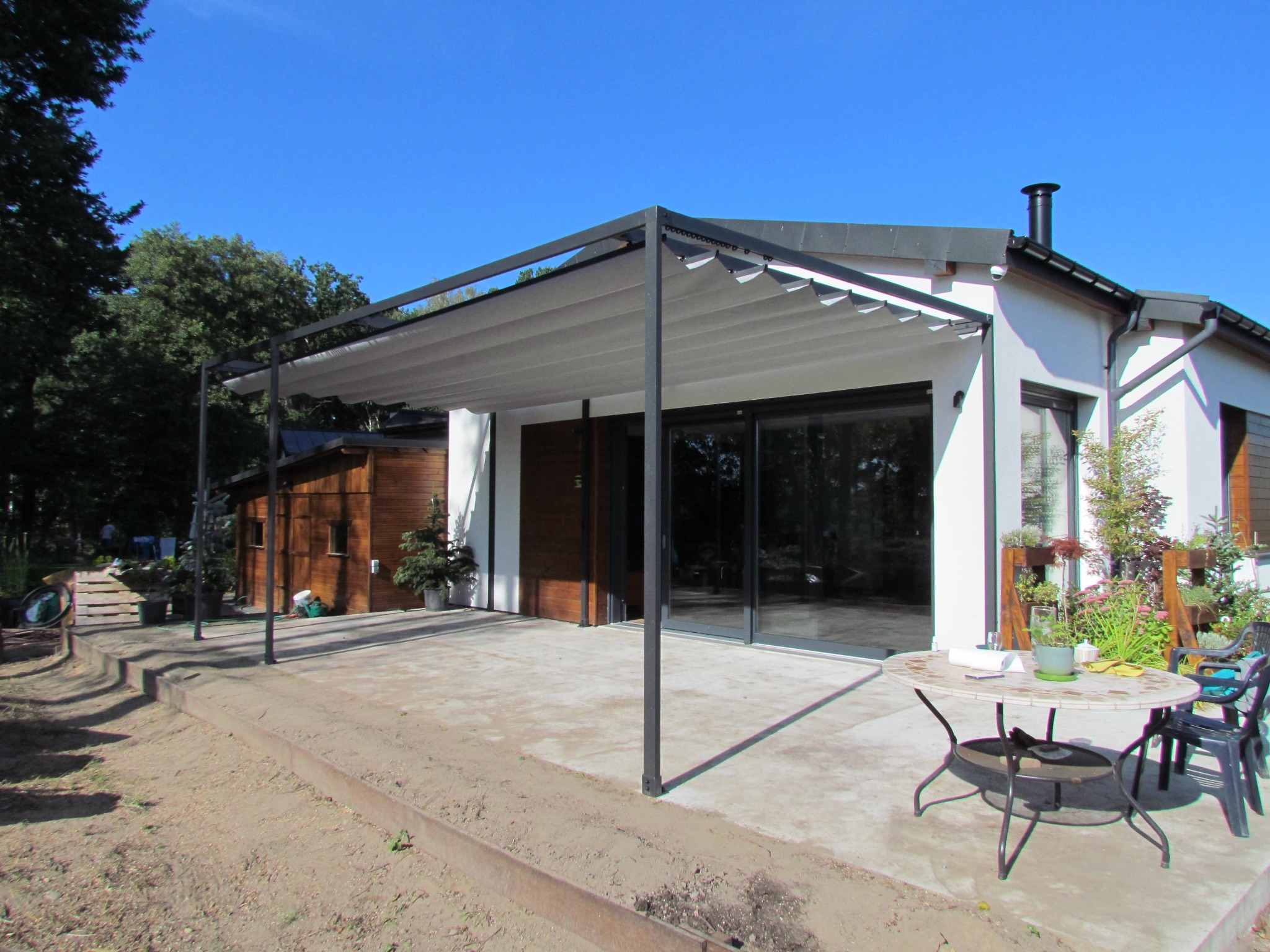 Nowoczesna pergola metalowa z rozsuwanym, jasnym zadaszeniem, przylegająca do białego domu z dużymi przesuwnymi oknami, na betonowym tarasie z okrągłym stolikiem i krzesłami.