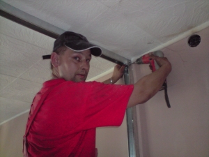 Mężczyzna w czerwonej koszulce i czapce w trakcie montażu metalowej konstrukcji sufitu podwieszanego za pomocą wkrętarki. Widoczne profile przy ścianie i suficie. Ściana w kolorze beżowym.