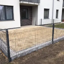 Nowo wyłożony, piaskowy taras z betonowych płyt, otoczony niskim, metalowym ogrodzeniem z drobnym, białym grysem po obwodzie, na tle białego budynku wielorodzinnego z ceglanym detalem.