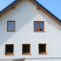 Usługi elewacyjne - Biała elewacja domu jednorodzinnego z drewnianymi oknami, widoczne worki z materiałami budowlanymi i fragment drabiny przy ścianie.