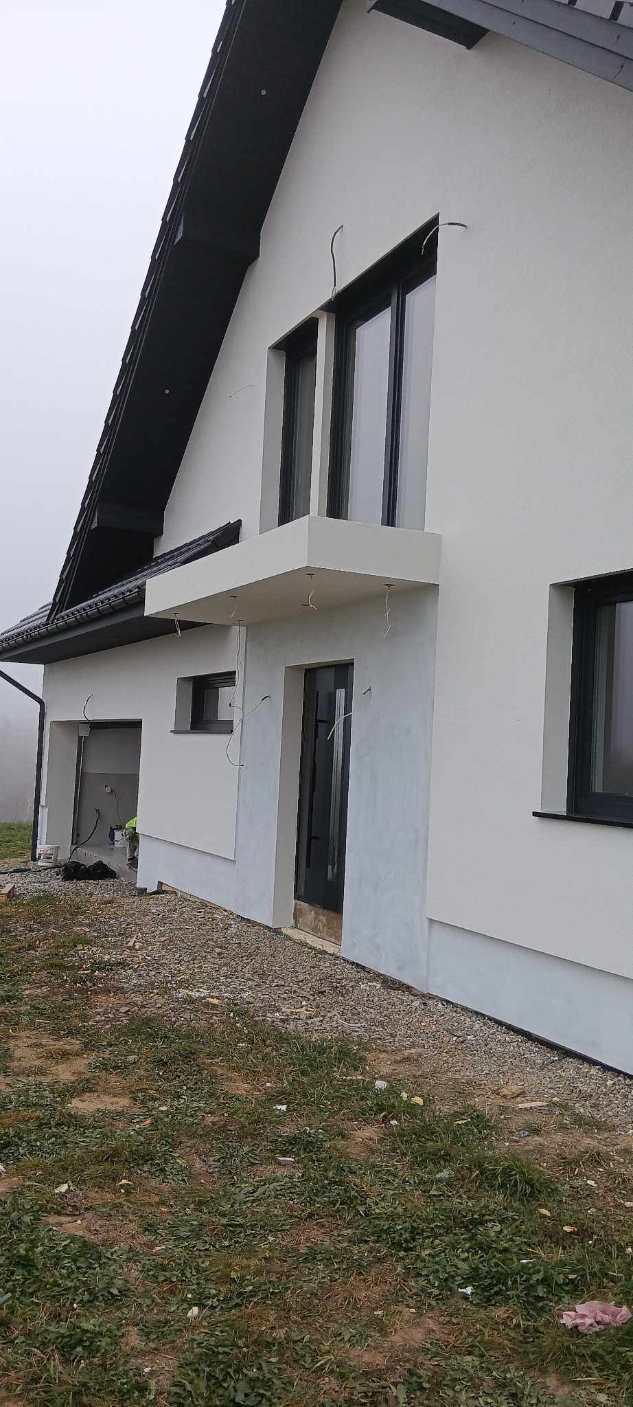 Nowoczesny dom jednorodzinny z białą elewacją w trakcie prac wykończeniowych, widoczne przewody elektryczne wystające ze ściany, szara podmurówka i czarne ramy okienne, teren wokół domu częściowo...
