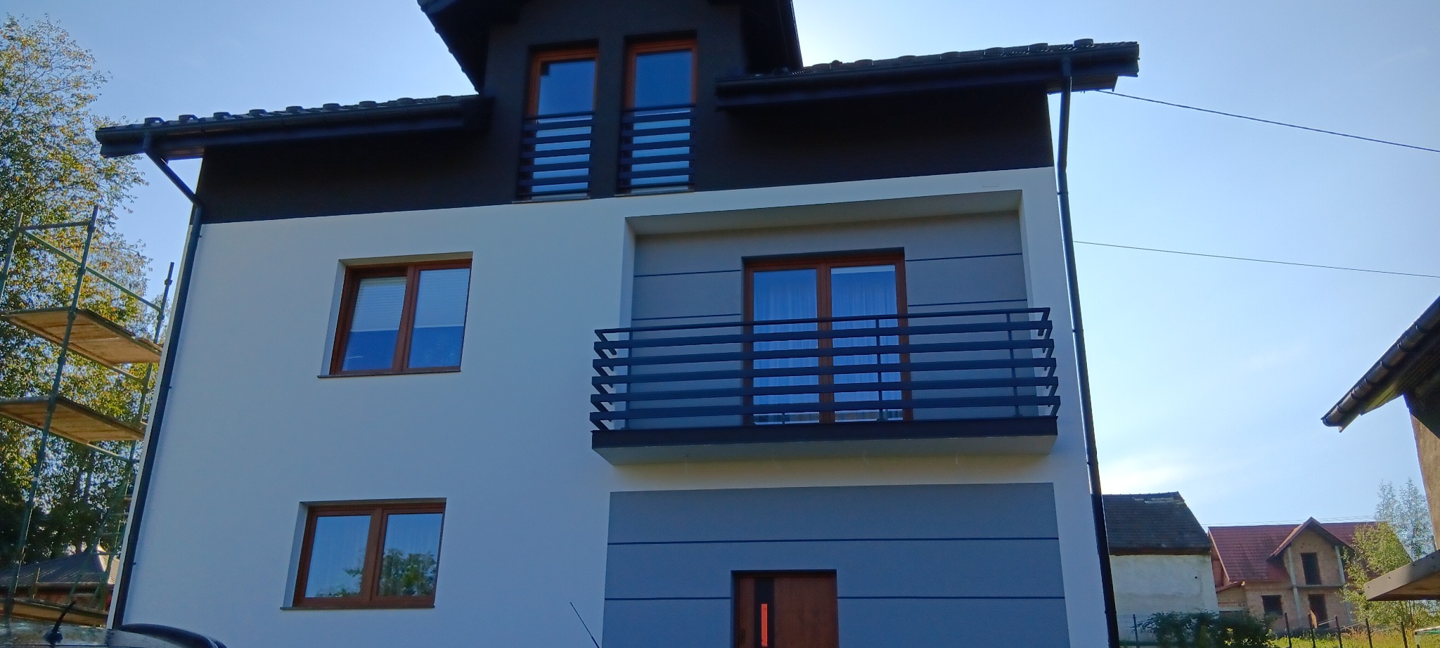 Dwukondygnacyjny dom z nową, dwukolorową elewacją w odcieniach bieli i szarości, z balkonem z metalową balustradą, widoczne rusztowanie po lewej stronie.