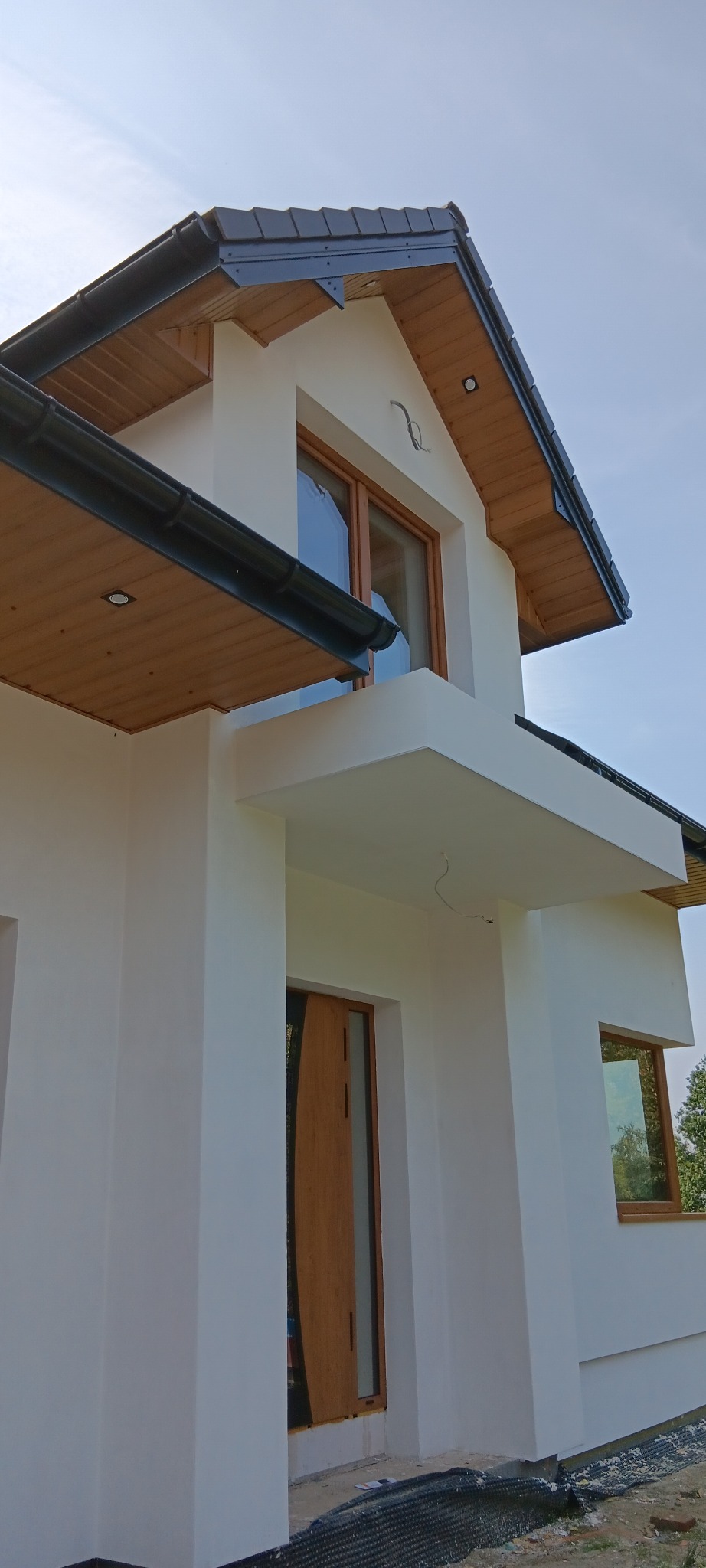 Nowoczesny dom z białą elewacją, drewnianymi oknami i czarnym dachem, widok z dołu na fragment budynku z widocznymi niedociągnięciami w wykończeniu wokół drzwi wejściowych.
