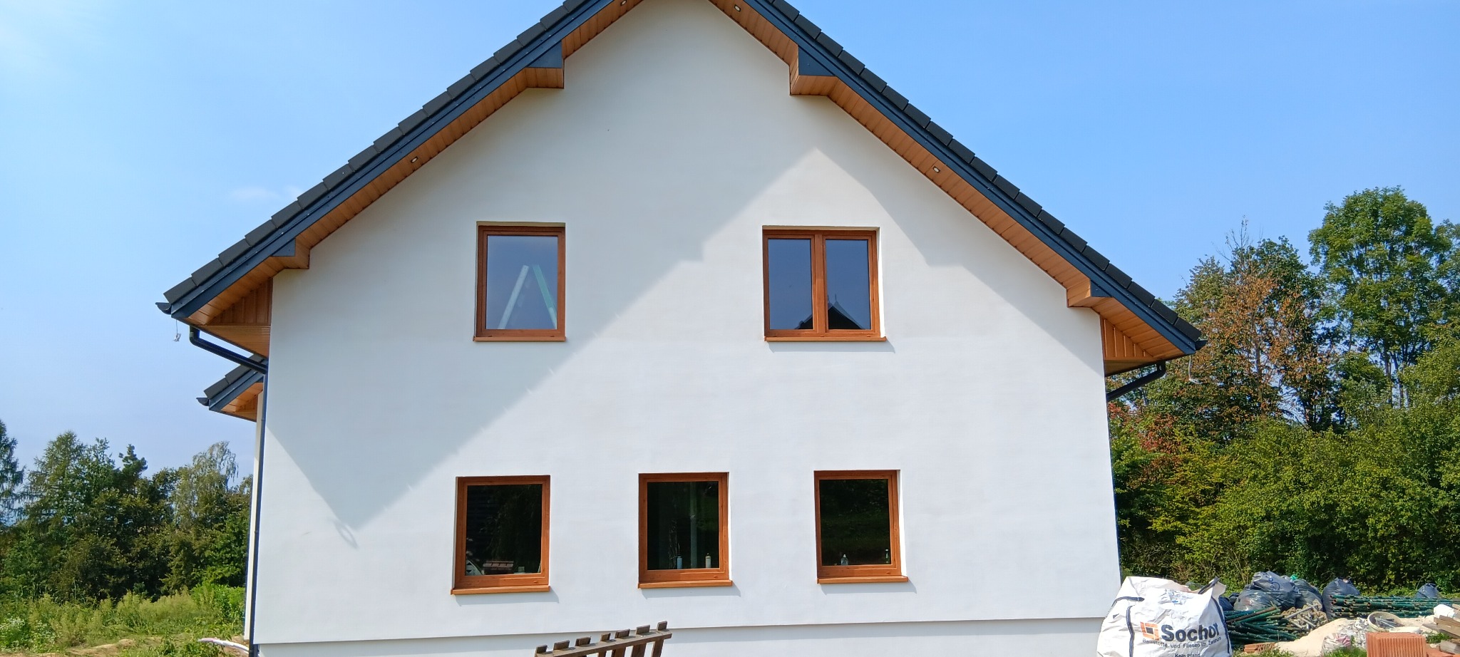 Biała elewacja domu jednorodzinnego z drewnianymi oknami, widoczne worki z materiałami budowlanymi i fragment drabiny przy ścianie.