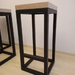 Dwa nowoczesne hokery barowe z czarną, metalową ramą i siedziskiem z jasnego drewna, ustawione na drewnianej podłodze przy białej ścianie.