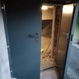Otwarta, metalowa brama wjazdowa w piwnicy, z widocznym wnętrzem pomieszczenia wypełnionego gruzem i oświetlonego żarówką.