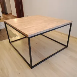 Kwadratowy stolik kawowy z blatem z jasnego drewna klejonego i czarną, metalową ramą w minimalistycznym stylu, ustawiony na panelach podłogowych w jasnym odcieniu drewna.