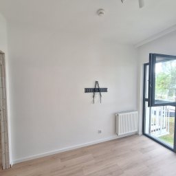 FIRMA USLUGOWA DARIUSZ WALKOWSKI - Puste, jasne pomieszczenie z panelami podłogowymi, grzejnikiem, uchwytem na telewizor na białej ścianie i otwartym oknem balkonowym.