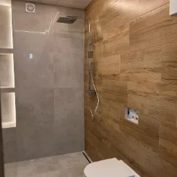 Nowoczesna łazienka z kabiną typu walk-in, wykończona płytkami imitującymi drewno i szarym gresem, podświetlane wnęki, wisząca toaleta. Minimalizm.