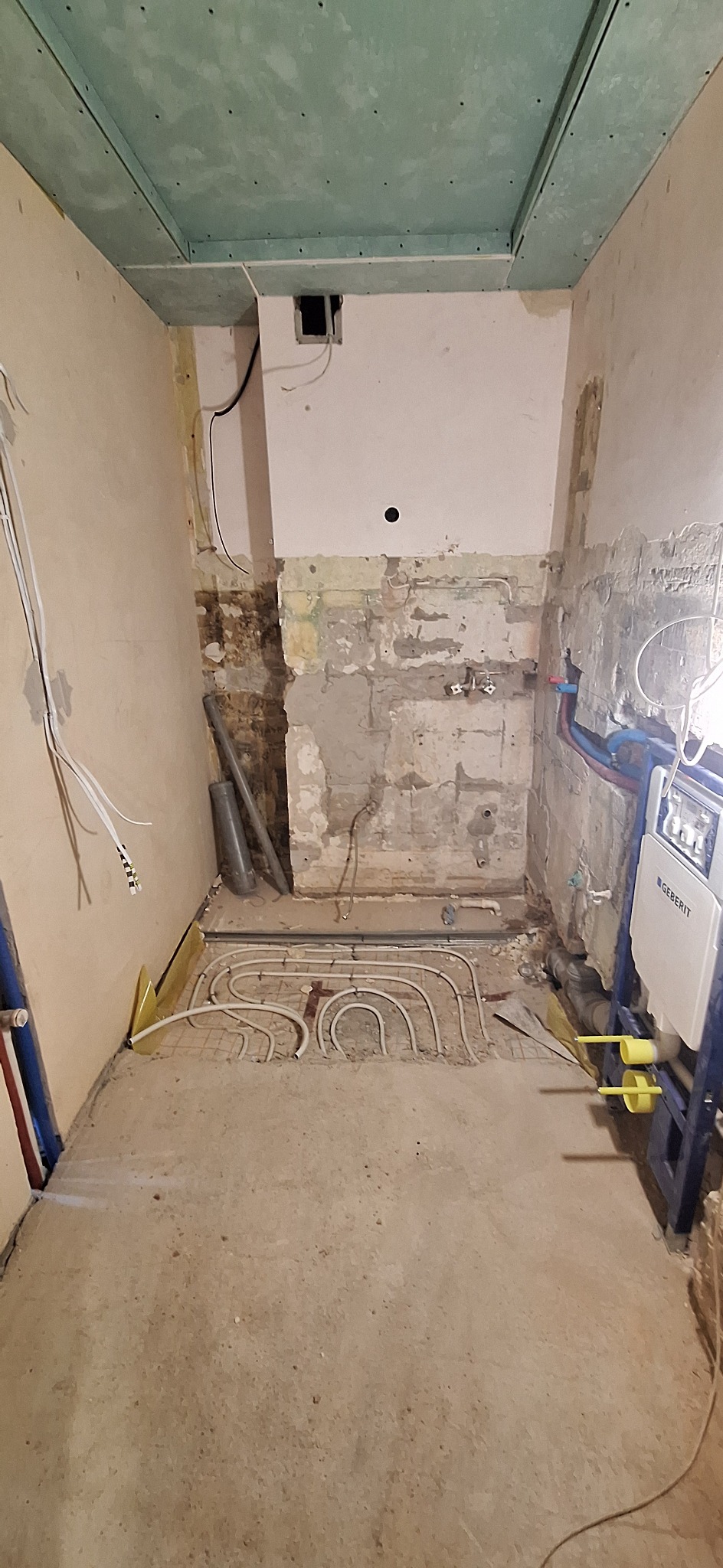 Łazienka w trakcie remontu: widoczne rury ogrzewania podłogowego, ściany z fragmentami starego tynku i zamontowany stelaż WC Geberit.