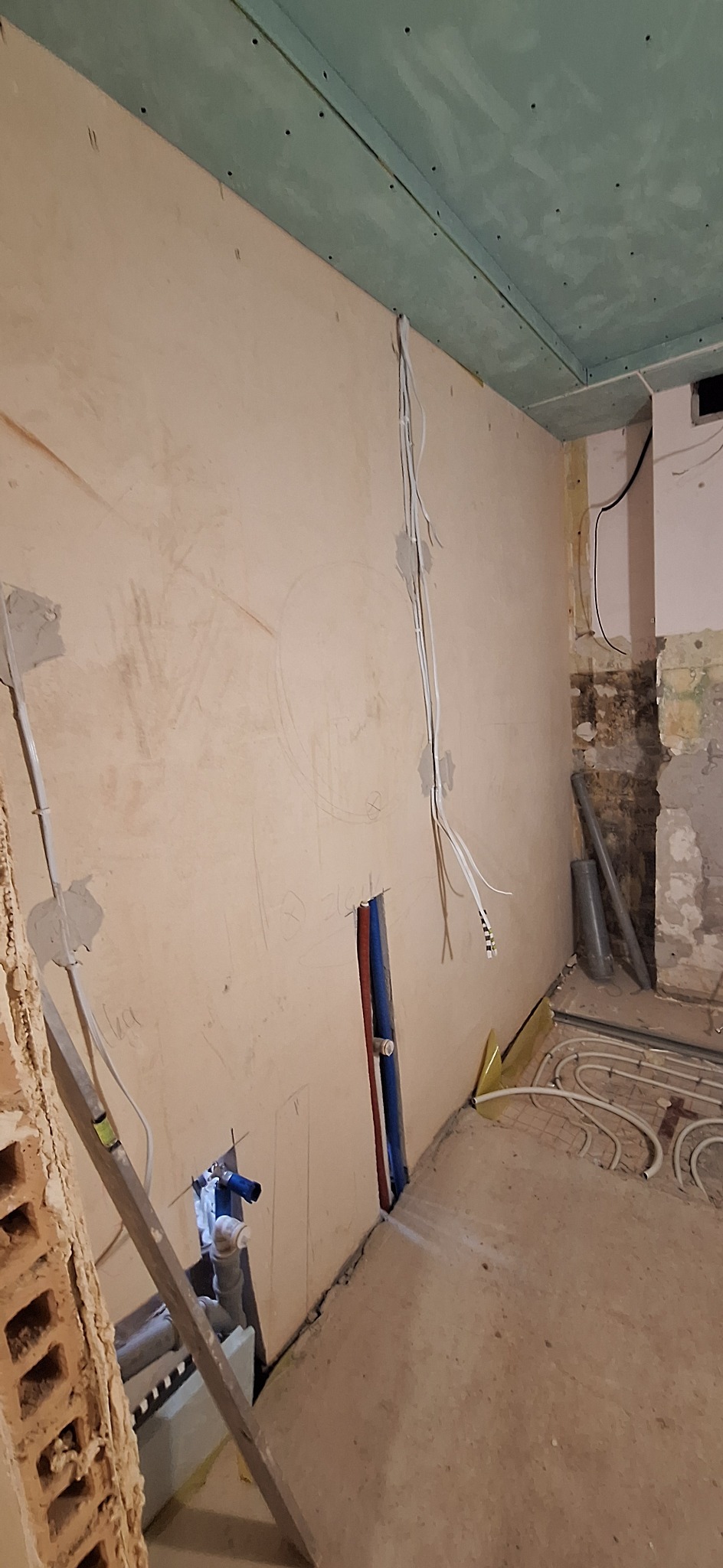 Ściana z instalacją elektryczną i hydrauliczną w trakcie remontu. Widoczne rury ogrzewania podłogowego i płyta kartonowo-gipsowa na suficie.