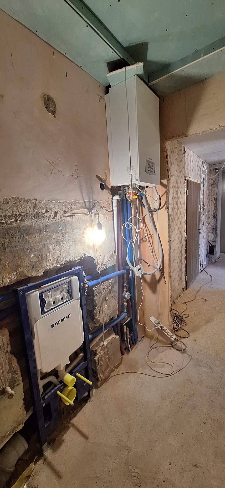 Remont łazienki w trakcie: zamontowany stelaż podtynkowy Geberit, widoczne rury instalacji wodnej i gazowej, surowy stan ścian i podłogi, instalacja elektryczna.