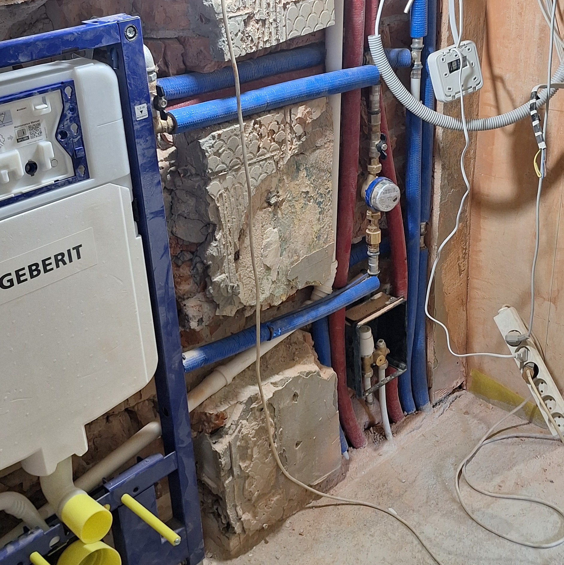 Montaż stelaża podtynkowego Geberit z instalacją wodną (rury PEX niebieskie i czerwone) na tle surowej ściany. Widoczny licznik wody i gniazdko elektryczne.