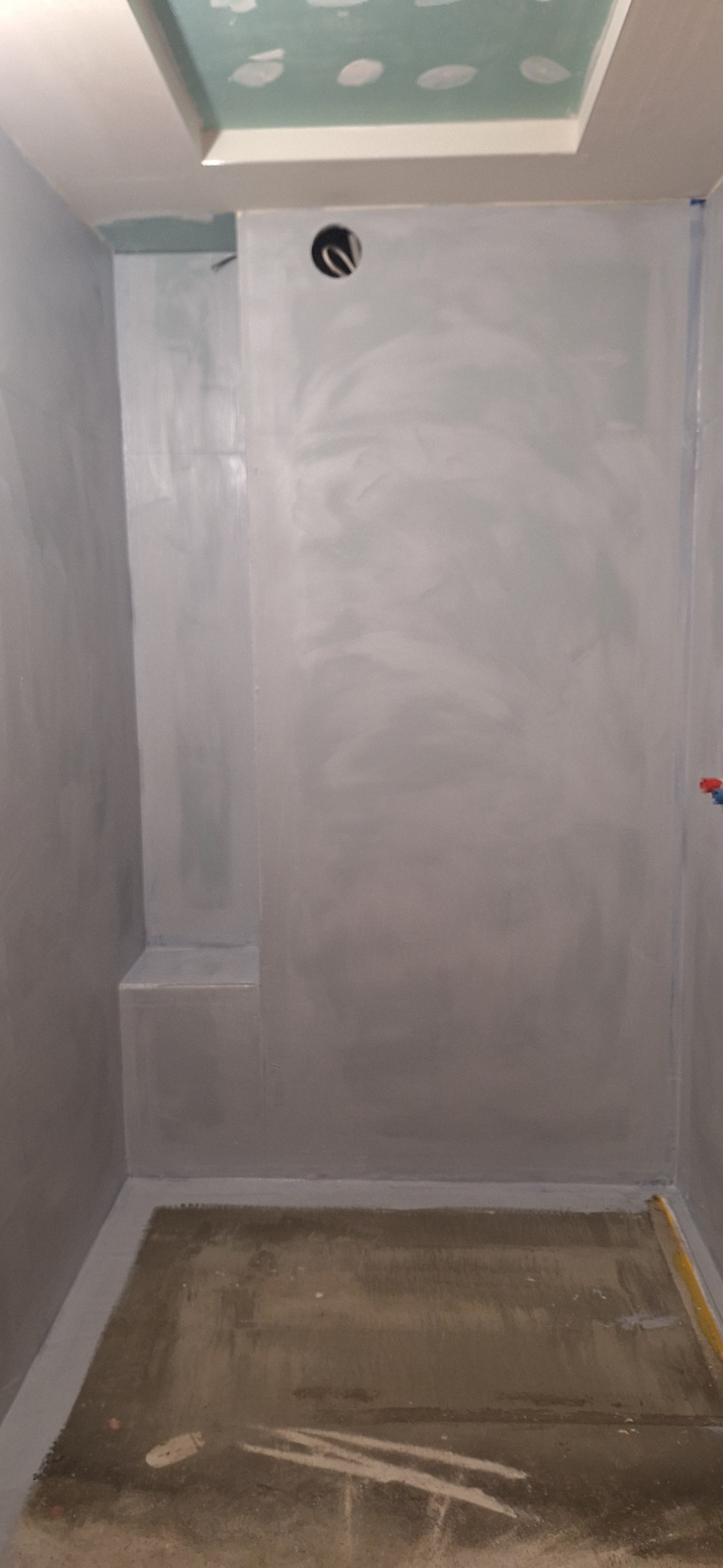 Przygotowana do dalszych prac ściana w łazience z wnęką, pokryta szarą hydroizolacją. Widoczny sufit z płyt gips-karton, otwór na oświetlenie i fragment instalacji wodnej.