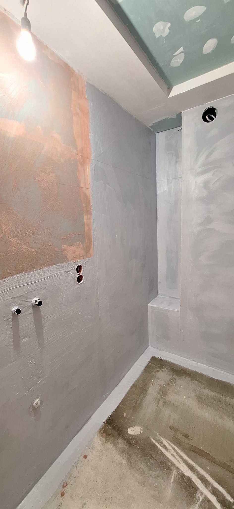 Surowa łazienka w szarościach: ściany po gruntowaniu, widoczne instalacje wodne i elektryczne. Przygotowanie do dalszych prac wykończeniowych.