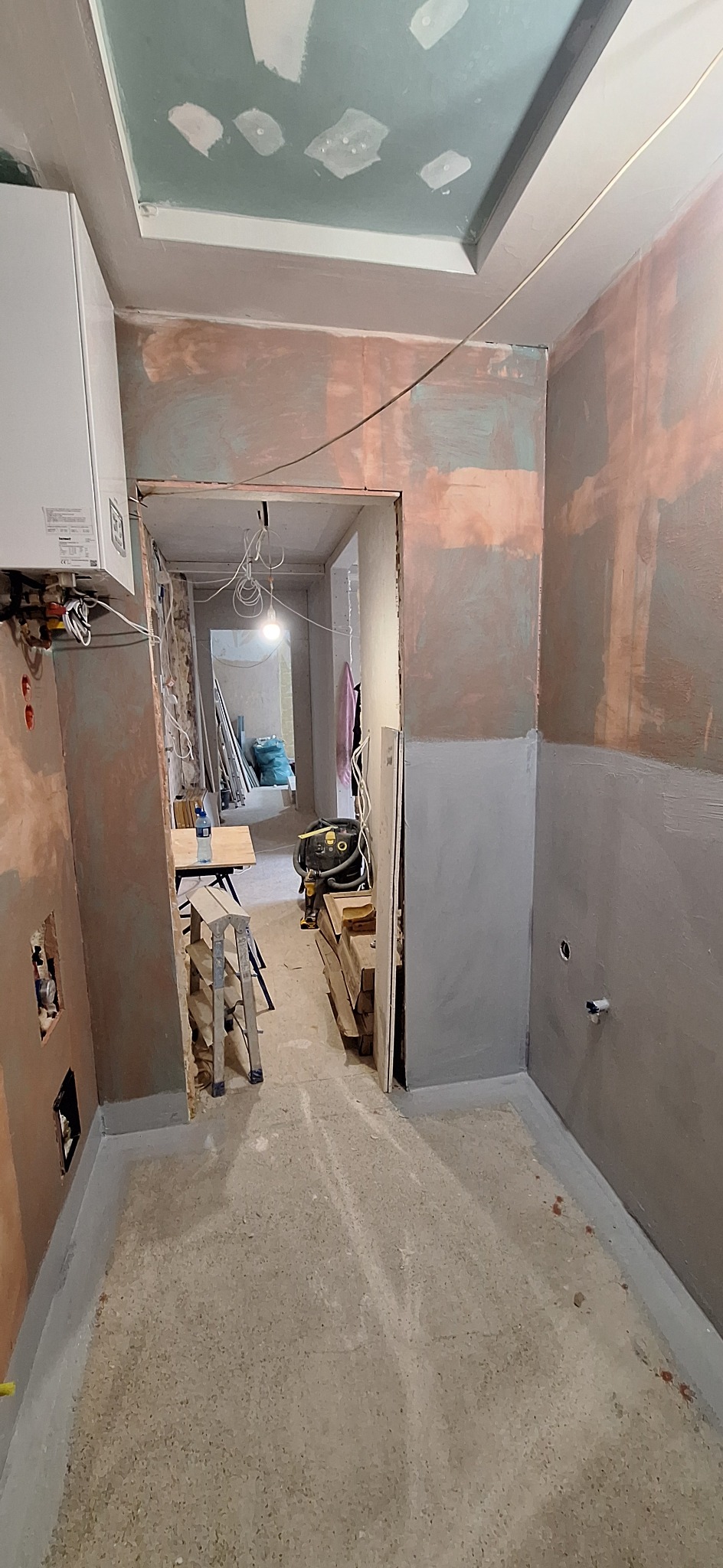 Wnętrze łazienki w trakcie remontu: surowe ściany z widocznymi śladami szpachlowania, przygotowane do dalszych prac wykończeniowych. Widoczne instalacje i narzędzia.