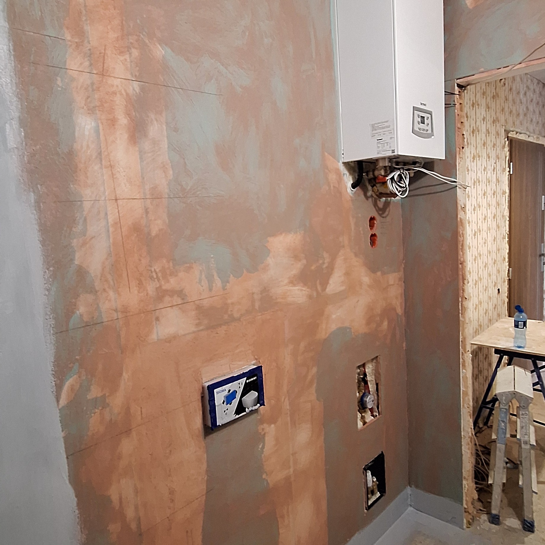 Ściana łazienki w trakcie remontu: widoczne instalacje, przygotowana powierzchnia pod płytki, piec gazowy na ścianie. Prace wykończeniowe w toku.
