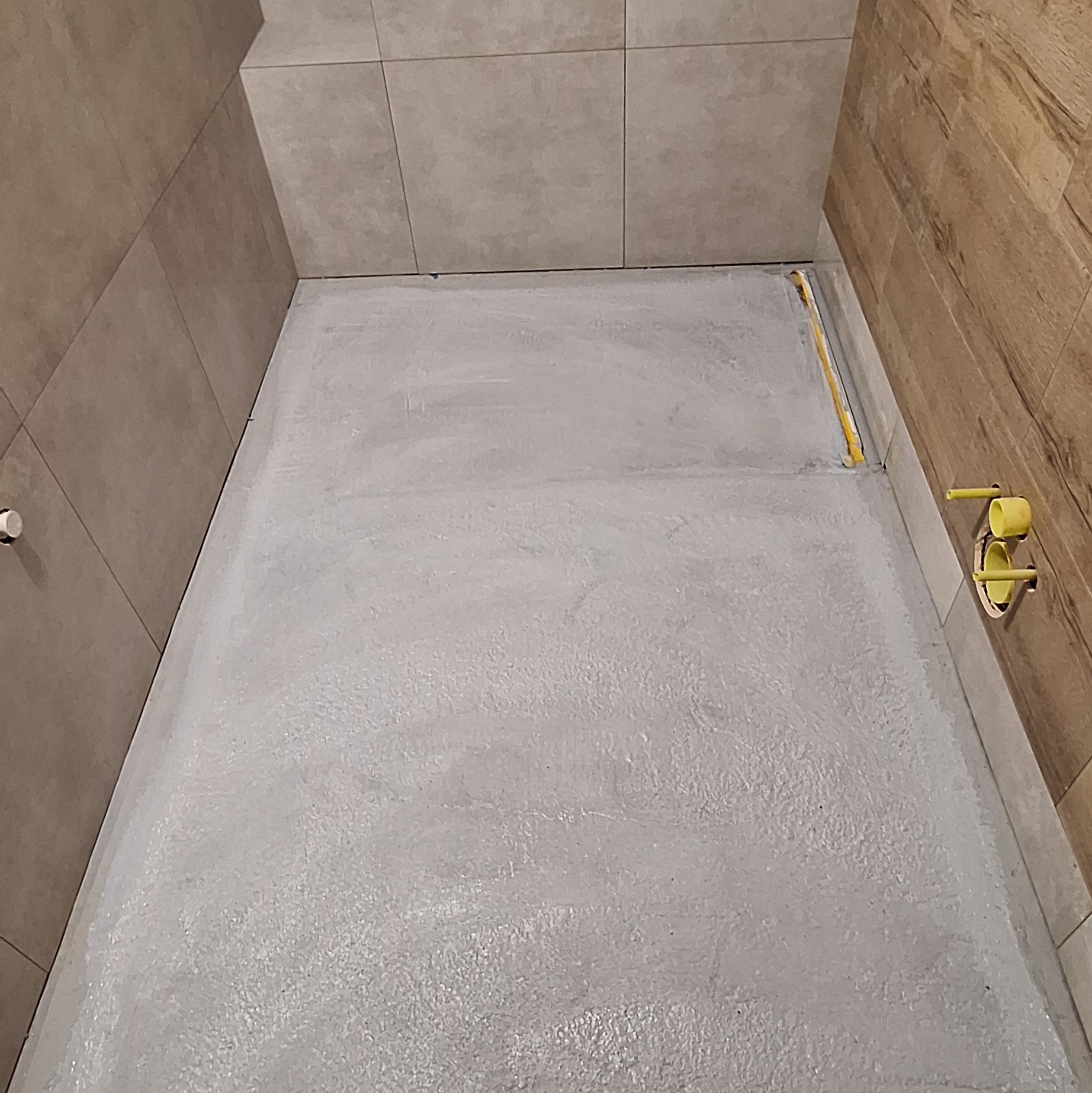 Przygotowanie podłoża pod prysznic: wylewka wyrównująca z hydroizolacją, widoczne rury odpływowe. Ściany wyłożone płytkami ceramicznymi w odcieniach szarości i drewna.