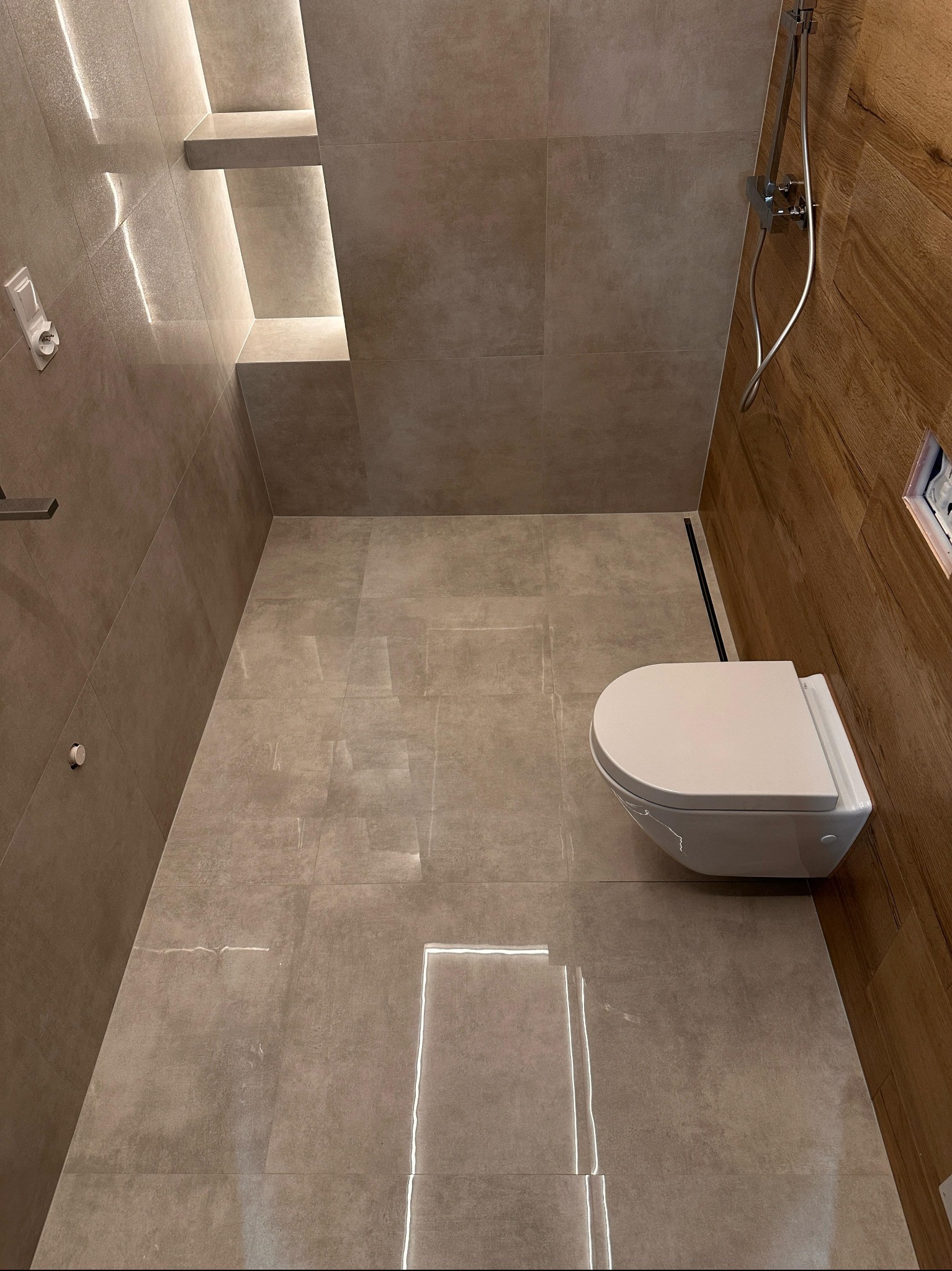 Nowoczesna łazienka w beżowych płytkach z drewnianym akcentem, podświetlane półki wnękowe i wisząca toaleta. Minimalistyczny design z odpływem liniowym.