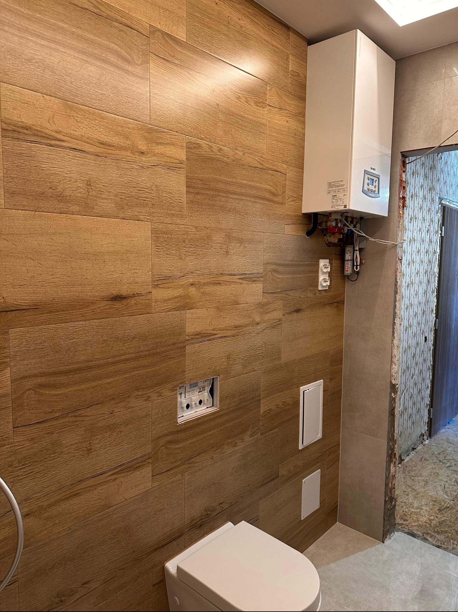Łazienka z drewnopodobnymi płytkami na ścianie, wisząca toaleta i piec gazowy. Widoczne białe klapy rewizyjne i otwór na stelaż podtynkowy.